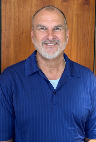 Smiling middle-aged man with short gray hair and stubble, wearing a blue polo shirt, posed against a wooden panel backdrop.