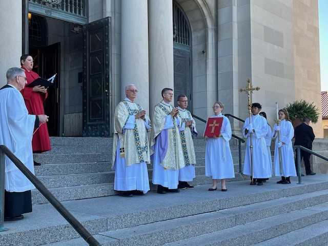 sfgjhgfd - St. Cecilia Cathedral Clergy and altar servers stand on church steps for a liturgical ceremony; priests in white and gold vestments, one holding a red liturgical book, others with a cross and sacred items.